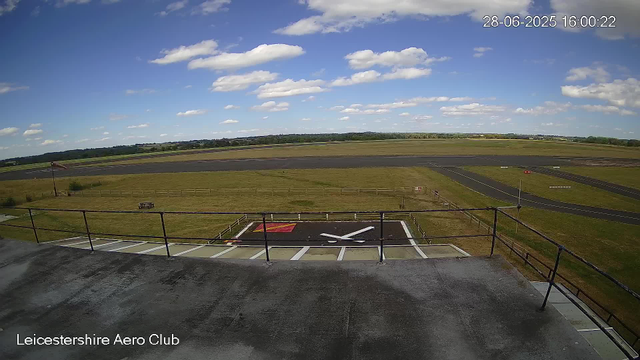A view from a webcam at Leicesterhire Aero Club showing an open tarmac area with a helipad marked by a white "H" painted on the ground. The sky is partly cloudy with fluffy white clouds scattered across a bright blue sky. In the distance, there are open fields and a runway, with a small flagpole and a banner visible near the helipad. The timestamp on the image indicates it was taken on June 28, 2025, at 4:00 PM.