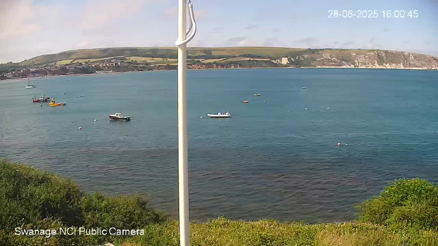 A tranquil coastal scene with a calm blue bay containing several small boats floating near the shore. In the background, there are green rolling hills with patches of trees, and white cliffs along the coastline. The sky is partly cloudy, with soft light suggesting late afternoon. A white flagpole is visible in the foreground, partially obstructing the view. The timestamp in the top right corner indicates the date and time as June 28, 2025, at 4:00 PM.