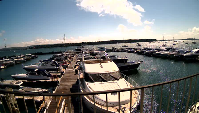 A marina with numerous boats docked along wooden piers, under a bright sky with some clouds. A large white yacht is prominently in the foreground, with other smaller boats lined up on either side. The water is shimmering, reflecting the sunlight, and a distant shoreline is visible on the horizon.