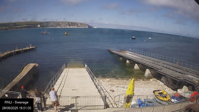 A sunny day at a coastal pier with people standing on the deck, some wearing hats. The pier extends over calm water, with a few boats floating nearby. In the background, there are green hills and white cliffs along the coast. The sky is partly cloudy. A yellow kayak and some other watercraft are stored on the shore below. There is a timestamp in the corner indicating the location as Swanage and the date as June 28, 2025.