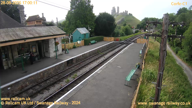 A small train platform with a stone building on the left, featuring a covered waiting area and adjacent flower baskets hanging from poles. There are green benches and a small green shed on the platform. Multiple railway tracks run through the scene, with a wooden fence and some greenery along the right side. An electrical pole with wires stands near the tracks on the right, and a narrow path runs parallel to the tracks through tall grass and trees. In the background, a hill with ruins or castle remains is visible atop it, surrounded by lush green trees. The date and time are displayed in the top left corner as "28-06-2025 17:00:11."