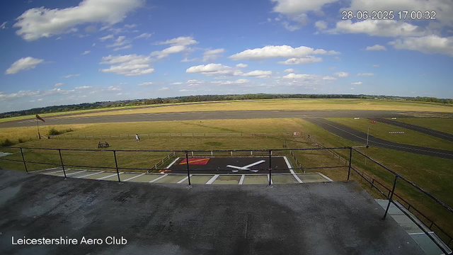 A view from a webcam at Leicestershire Aero Club shows a clear sky with scattered white clouds. In the foreground, a helicopter landing pad with a large white helicopter symbol on a black background is visible, surrounded by a metal railing. To the left, there is a red windsock on a pole, and a wooden bench with a person seated nearby. A person is walking along a fence line, and another individual is standing further back. Beyond the grassy areas, the runway extends into the distance with a few small signs, and the landscape is flat with fields and trees on the horizon. The timestamp indicates the date and time as June 28, 2025, at 17:00:32.