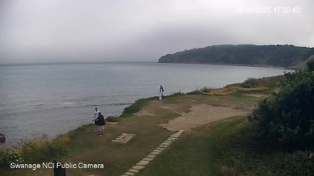 A view of a shoreline with three people standing near the water's edge, two of whom are wearing white clothing and one in dark clothing with a cane. The shoreline has a grassy and sandy area with a small stone path leading to the water. In the background, there is a large body of water extending to a distant, tree-covered headland. The sky appears overcast, and the date and time are displayed in the top right corner. The bottom left corner has text indicating it is from a "Swanage NCI Public Camera."