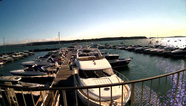 A marina filled with numerous boats and yachts docked along wooden piers, with calm water reflecting the sky and vessels. The horizon shows a distant landmass under a clear sky during sunset or late afternoon.