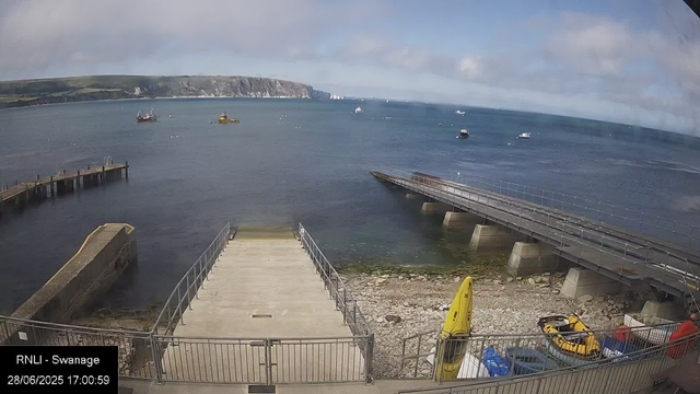 A view of a seaside scene with a dock extending into the water, boats anchored in the harbor, and a cliff in the distance under a partly cloudy sky. There are some vehicles, equipment, and a yellow life raft on the shore in the foreground. The timestamp indicates the photo was taken on June 28, 2025, at 17:00:59.