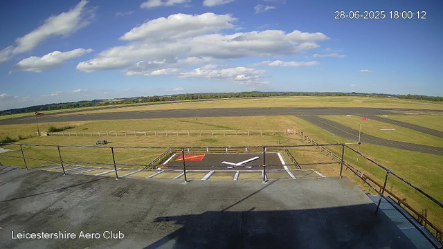 An outdoor view from a webcam at Leicestershire Aero Club shows a bright sky with scattered white clouds. In the foreground, there is a helipad with a large white "H" and red markings, surrounded by a metal fence. The tarmac and grassy areas are visible, with the runway in the background extending across the scene. The image's timestamp reads "28-06-2025 18:00:12".