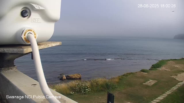 A webcam view shows a calm ocean shoreline under a hazy sky. In the foreground, part of the camera's white control device is visible, mounted on a concrete ledge. To the right, there is a grassy area with some patches of paving stones and pathways. The ocean has gentle waves, and a small rock formation is just offshore. In the background, a distant landmass or headland is faintly visible through the mist. The timestamp indicates the date as June 28, 2025, at 6:00 PM. The image is labeled "Swanage NCI Public Camera."