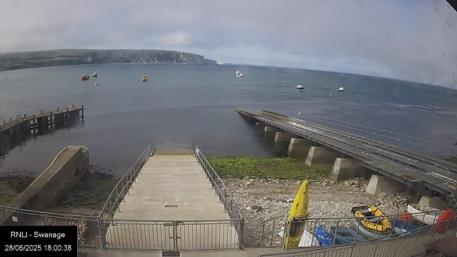 A view of a waterfront with a concrete pier extending into the water and moored boats scattered across the bay. In the foreground, there is a small area with a fence, a yellow rescue boat, and some equipment. A large wooded headland is visible in the background across the water under a partly cloudy sky. The timestamp indicates the date as June 28, 2025, at 6:00 PM.