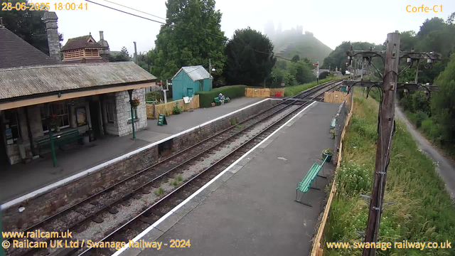 A small train station platform with a stone building featuring a pitched roof on the left. There are two green benches and a few small potted plants. One person with a riding helmet and riding gear is seated on a bench near a small green shed. To the right, a set of railway tracks runs through the scene, with a yellow safety barrier across the track in the distance. Tall green trees and hills are visible in the background, shrouded in a slight mist. An old wooden utility pole with multiple wires stands on the right side, and there is a narrow path with tall grass running parallel to the tracks on the far right. The scene is captured on an overcast day, and the timestamp shows it was taken on June 28, 2025, at 18:00.