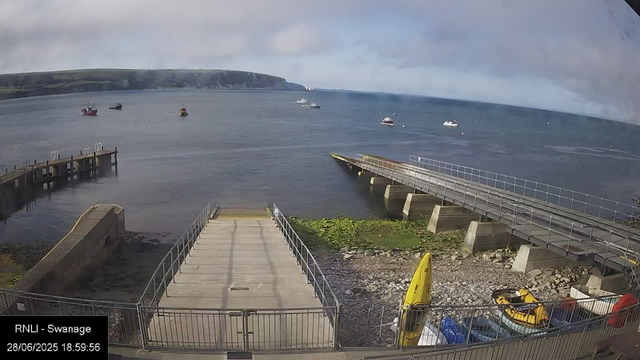 A view of a harbor with a concrete dock extending into the water, surrounded by metal railings. Several boats are anchored in the water, with a backdrop of cliffs and cloudy sky. There are a few yellow and red kayaks or small boats stored near the railing, and the time stamp indicates the photo was taken on June 28, 2025, at 6:59 PM at RNLI Swannage.