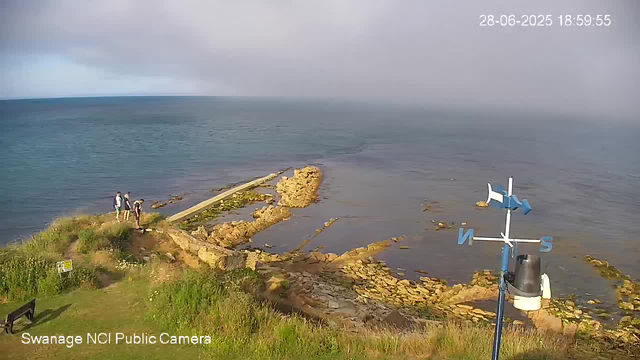 A panoramic view of a coastal scene with a rocky shoreline extending into the water, under a cloudy sky. Three people stand near the edge of the rocks, looking toward the sea. In the foreground, there is an area with green grass and bushes, along with a bench and a sign. To the right, a weather vane with directional indicators (N, S, E, W) is mounted on a pole. The timestamp in the upper right corner shows the date as June 28, 2025, and the time as 6:59:55 PM. The camera is identified as the "Swanage NCI Public Camera."
