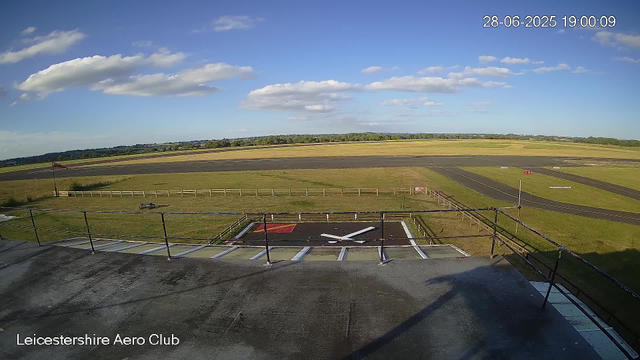 A wide view of an outdoor airfield on a sunny day with a partly cloudy sky. In the foreground, there is a rooftop with a large clock painted on it, showing the time as approximately 4:46. The airfield has a runway, some grassy areas, and a small fence, with a windsock visible on the left side. The digital timestamp indicates the date as June 28, 2025, and the time as 19:00:09. The bottom left corner displays the text "Leicestershire Aero Club."