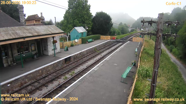 A small train station platform with a stone building on the left, decorated with hanging flower baskets. There are three green benches along the platform, a sign that says "WAY OUT", and a small turquoise shed. Overhead, power lines stretch across the scene. To the right, a grassy area with wildflowers runs beside the railway tracks, which curve away into a misty, green, hilly landscape. The scene appears early evening, with fog present in the background.