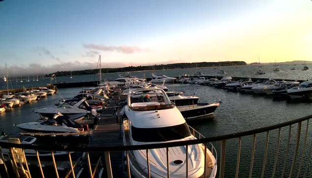 A marina filled with numerous boats and yachts docked along wooden piers, with the calm water reflecting the evening sky. In the background, there is a low horizon with trees and a few distant boats, under a partly cloudy sky illuminated by the setting sun.