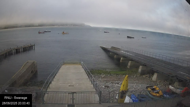 A view of a harbor with several boats floating on calm water under a cloudy sky. In the foreground, there is a concrete pier with metal railings, and an adjacent ramp leading into the water, with some green moss or algae on the rocks below. On the right side, there is a yellow kayak or small boat, along with various containers and equipment. The background features a shoreline with low hills, shrouded in mist or fog.
