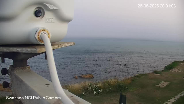 A view of the ocean with a rocky outcrop in the water, taken from a seaside location. A surveillance camera is visible at the top left corner of the image, with a cable hanging down from it. The timestamp in the top right corner shows the date and time as 28-06-2025 at 20:01:03. The scene includes a grassy area with stone pavers on the right side and some plants near the shoreline.