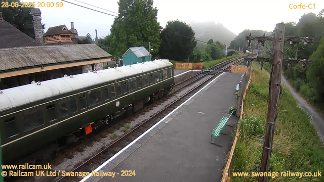 A vintage green train with a white roof is parked at a small rural station platform surrounded by trees and greenery. The platform has a couple of green benches, some potted plants, and a steady mist in the background. Overhead, there are electrical lines supported by wooden poles. To the left, there are buildings with tiled roofs and chimneys. The scene appears to be early evening or late afternoon, as indicated by the timestamp. The area is quiet and scenic, with a narrow path running alongside the tracks.