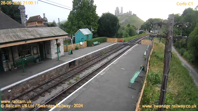 A small train station platform with a stone building on the left, green benches, and a small teal shed in the background. Railroad tracks run through the scene, curving to the right, with a wooden utility pole and wires overhead. There is greenery on both sides, including trees and bushes, with a hill and castle ruins visible in the distance. The photo was taken in the early evening, as indicated by the timestamp.
