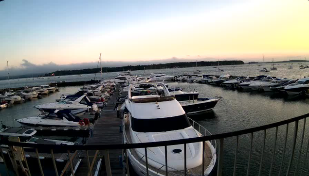 A marina filled with various boats and yachts docked along piers on calm water during sunset, with a distant shoreline and a few clouds in the sky.