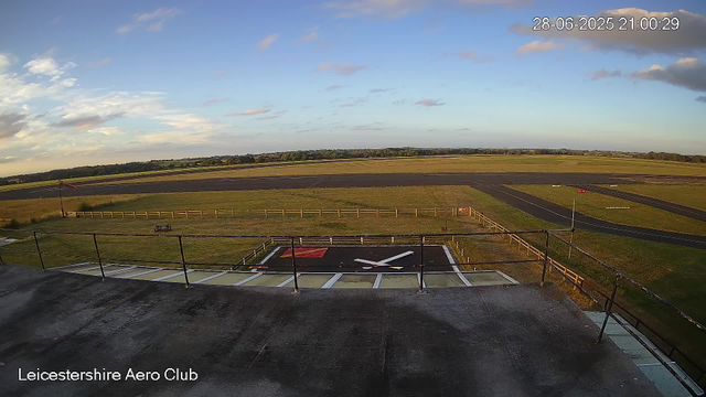 An outdoor scene from a webcam at Leicesterhire Aero Club shows a large open sky with a few scattered clouds. In the foreground, there is a large helipad with a white "H" marking and a red rectangle, surrounded by a black railing. To the left, a small flagpole with a red flag flies. Beyond the helipad, there are green grassy fields and a winding paved path. In the distance, a low horizon line separates the fields from the sky, and the time stamp in the upper right corner indicates the date is June 28, 2025, at 9:00 PM. The bottom left corner displays the text "Leicestershire Aero Club."