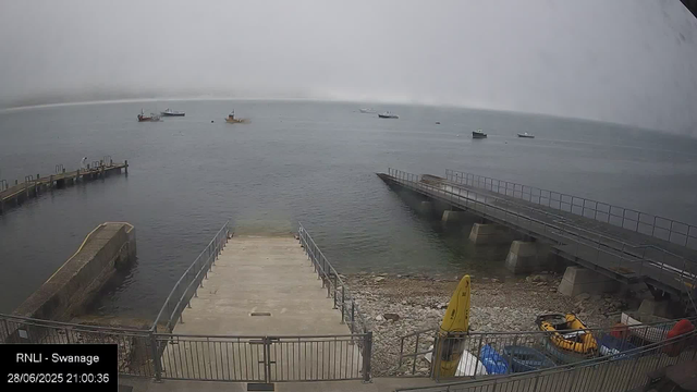 A view of a harbor on a foggy day, with several boats floating on calm water in the distance. In the foreground, there are two concrete piers extending into the water, with metal railings along their edges. To the right, a sloped walkway with railings leads down to the rocky shoreline, which has some equipment including a yellow surf rescue board, a yellow life jacket, and other items. The scene is overcast and misty, obscuring the horizon.