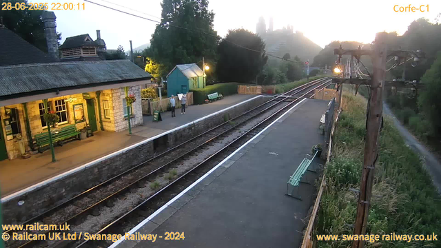A small railway station platform during early evening with two people standing near a green bench and a small blue building. The station building is made of stone with green doors and flower baskets hanging outside. There are benches along the platform and a wooden fence between the platform and the train tracks. In the background, a hillside with trees and some faintly visible buildings or structures under a partly cloudy sky. A wooden utility pole with electrical wires is on the right side of the platform. The lighting is warm, indicating sunset nearby.