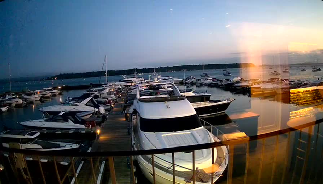A marina filled with various boats and yachts docked along wooden piers during sunset, with calm waters reflecting the sky and vessels. The horizon shows a distant shoreline under a sky fading from blue to orange and pink hues.