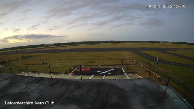 A wide view of an airfield taken from a webcam at the Leicester Aero Club on June 28, 2025, at 10:00 PM. The foreground shows a helicopter landing pad with large white and red markings. Beyond the pad, there is a grassy area enclosed by a wooden fence, with a small windsock on a pole to the left. The runways extend into the distance with clear markings, surrounded by grassy fields under a partly cloudy sky during dusk.