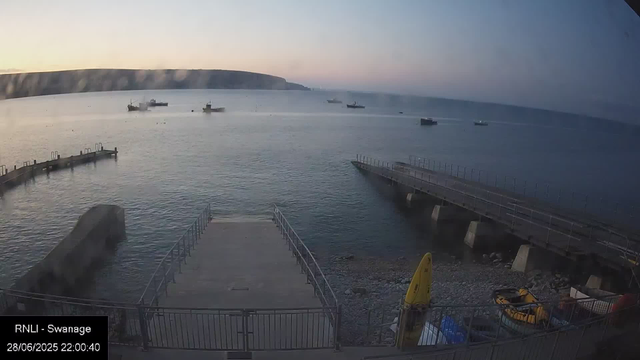 A view of a harbor at dusk with calm water and several small boats floating in the distance. In the foreground, there are two concrete piers or breakwaters extending into the water, with a fenced area containing a yellow caution sign and some equipment. The sky is a gradient from light to dark, indicating early evening. The scene appears peaceful with soft natural light.