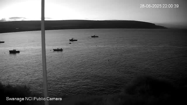 A black-and-white image of a body of water with several small boats floating on the surface. A distant shoreline or hill is visible in the background. The sky appears mostly clear with a few clouds, and the time and date are displayed in the top right corner as "28-06-2025 22:00:39". The bottom left corner has the text "Swanage NCI Public Camera."