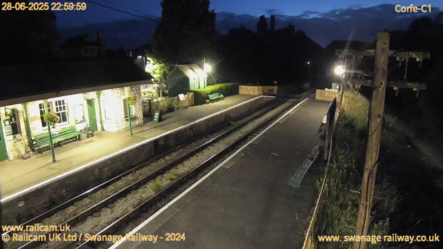 A quiet train station platform at night, illuminated by a few overhead lamps. The platform has a green bench, a small shelter, and some hanging flower baskets outside a stone building with green doors and window frames. There are a few signs, including one indicating "WAY OUT." The railway tracks run parallel to the platform, with a wooden fence and some benches along the right side. The sky is dark, and mountains are visible in the distance. A large wooden utility pole with wires is on the right side of the image. The timestamp indicates it is 10:59 PM on June 28, 2025.