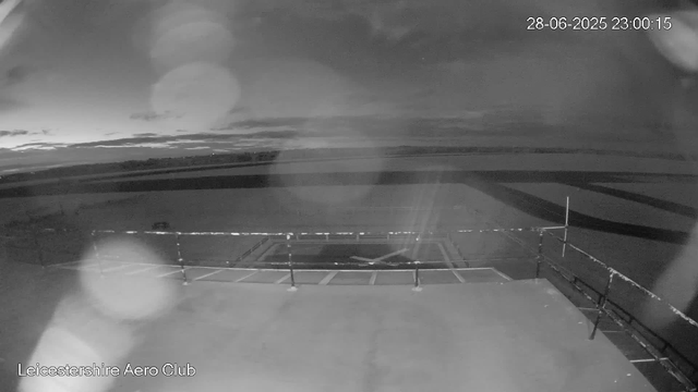A black-and-white image from a webcam showing an outdoor area at night, with clouds in the sky and a distant horizon. The ground has parking spaces and a metal railing along the edge. The date and time stamp in the upper right corner reads "28-06-2025 23:00:15," and the bottom left corner displays "Leicestershire Aero Club."