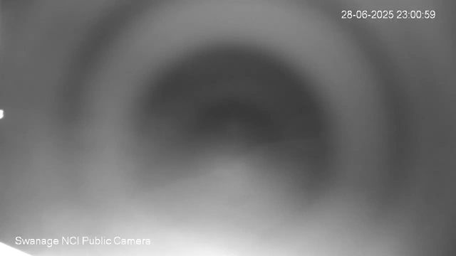 A blurry, black-and-white close-up image of a circular object with a small opening in the center, taken from a webcam. The timestamp in the upper right corner indicates the date and time as 28-06-2025 23:00:59. The bottom left corner shows the label "Swanage NCI Public Camera."