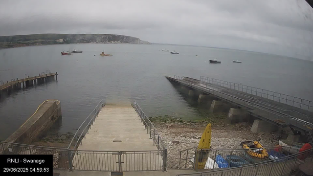 A view of the harbor at Swange with several boats anchored in the water, a dock extending into the sea, and a cloudy sky overhead. There is a concrete and metal railing on a small platform at the water's edge, with a yellow lifeguard float and some equipment nearby on the rocky shore. The landscape includes cliffs in the background under an overcast sky.