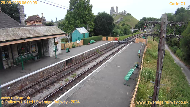 A small train station platform with a stone building on the left, featuring a sloped, tiled roof and hanging flower baskets. Green benches are spaced along the platform. To the right, there's a tall wooden utility pole with electrical wires. Behind the platform, there is a green shed and a neatly trimmed hedge. In the background, a grassy hill with a castle ruin on top is visible. The scene is outdoors with trees and greenery, and the sky appears clear. The image includes timestamp and details about the railway at the top and bottom of the image.