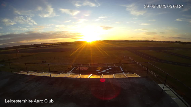 A sunrise over an airfield with the Leicester Aero Club logo at the bottom. The sky is partly cloudy with the sun just above the horizon, casting a warm glow and long rays across the landscape. The foreground shows a platform with a windsock and a helicopter landing pad. The airfield has grassy areas, runways, and some small structures in the distance. The photo is timestamped 29-06-2025 at 05:00:21.