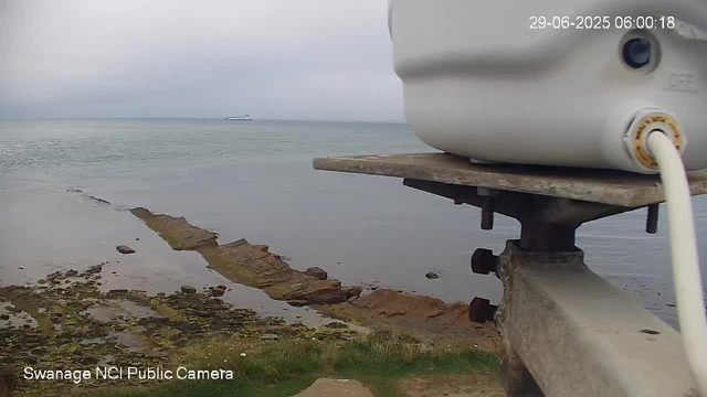 A view of a shoreline with calm water and a large ship in the distance. In the foreground, there is a close-up of a weathered wooden and metal structure with a white device attached to it, connected by a white cable. The sky is overcast, creating a grayish atmosphere. The bottom left corner has text that reads "Swanage NCI Public Camera," and the top right corner shows a timestamp of "29-06-2025 06:00:18."