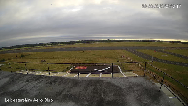 A wide view of an airport runway and grassy fields under a cloudy sky. In the foreground, a helipad with a white letter "H" and a red and white sign. A small flag and a bench are also visible on the grass area. The horizon shows distant trees and fields. The timestamp reads "29-06-2025 06:00:17," and the location is indicated as "Leicestershire Aero Club."