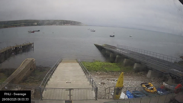 A view of a cloudy sky over a calm body of water with several boats floating; a concrete pier extends into the water, with a small ramp in the foreground leading to a fenced area containing a yellow kayak, a life ring, and some supplies. The landscape includes green hills in the distance. The timestamp shows it is early morning on June 29, 2025.