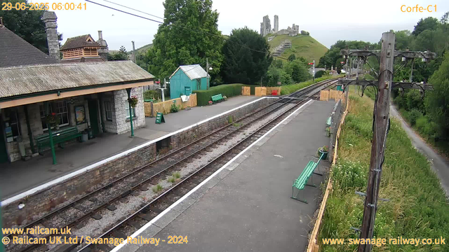 A small train station platform with a stone building on the left, featuring a green bench and hanging flower baskets. There are multiple green benches along the platform. Potted plants are placed next to the benches, and a sign indicates "Way Out." A wooden fence with yellow and green panels borders the platform, separating it from lush green trees and grass. Overhead, power lines run parallel to the tracks. In the background, there is a hill with ruins and a winding path leading up to them. To the right, there is a row of wooden utility poles with electrical wires, and a dirt path runs alongside a grassy area with wildflowers. The overall scene appears to be early morning, as indicated by the timestamp.