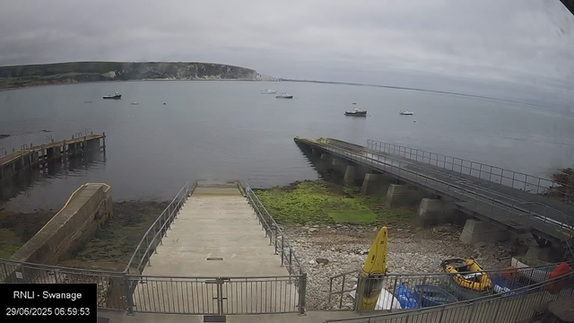A view of a harbor with several boats floating on calm water under a cloudy sky. In the foreground, there is a concrete pier and a ramp leading into the water. To the right, a metal bridge extends over water with mossy green patches underneath. There is a small fenced area at the bottom containing a yellow safety sign, a yellow inflatable boat, and some other equipment. A clock in the bottom left corner indicates the time as 06:59:53 on June 29, 2025. The location label reads "RNLI - Swanage."