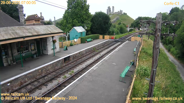 A small train station platform with a stone building on the left that has a sloped roof and green benches underneath the overhang. There are flower baskets hanging from the building, and a sign that says "WAY OUT" near the edge of the platform. To the right, there are also green benches and flower pots along a yellow fence. The railway tracks run down the middle of the image, with a grassy area and a path on the far right. In the background, there is a hill with ruins of an old castle on top, surrounded by trees and greenery. A wooden utility pole stands on the right side of the platform with some wires overhead. The sky is overcast. The timestamp indicates the image was captured on June 29, 2025, at 7:00 am.