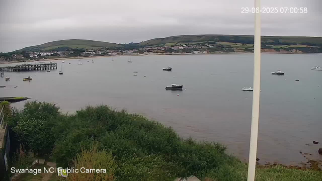 A view of a calm bay with several small boats anchored on the water. In the background, there are green hills and a small town along the shoreline. The sky is overcast, and there is a dock extending into the water on the left side. The foreground features green bushes and trees. The timestamp in the upper right corner indicates it is early morning.