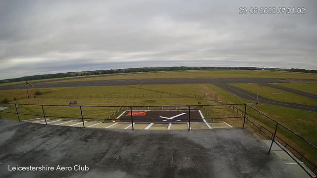 An overcast sky at Leicestershire Aero Club, showing a view of the runway and surrounding grassy areas. The photo is taken from a raised platform or balcony with a metal railing in the foreground. The grass area has a marked helicopter landing pad with a white 'H' and some signage. The runway extends into the distance, bordered by fields and trees under a cloudy sky. The timestamp in the upper right corner reads "29-06-2025 07:01:02".