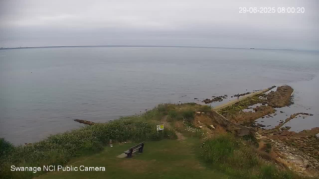 A wide view of a coastal area with calm water under an overcast sky. In the foreground, there is a grassy area with a bench facing the water, some bushes, and a small sign. Rocks extend into the sea, forming a narrow path or jetty. The date and time are displayed in the top right corner.