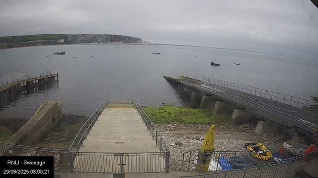A view of a small harbor with concrete and wooden piers extending into calm water under a cloudy sky. Several boats are anchored in the distance, and a grassy, rocky shoreline is visible in the foreground. On the lower right, there is a yellow kayak and some water bottles, with an area enclosed by a metal fence. The landscape in the background features green cliffs and coastal terrain. The image is timestamped "29/06/2025 08:00:21" and labeled "RNLI - Swanage."