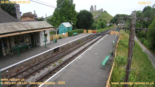 A small train station platform with green benches, a green shed, and decorative flowers. There are railway tracks running through the station, with a city castle visible in the background. The scene is surrounded by lush green trees and grassy hills. The sky is overcast. The timestamp on the image reads "29-06-2025 08:00:23."