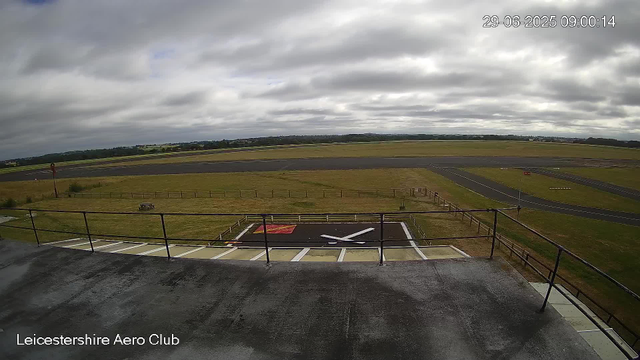 An overcast sky with thick, gray clouds above an empty runway at Leicestershire Aero Club. The takeoff/landing area is visible in the distance, surrounded by grassy fields. In the foreground, the rooftop of a building with a metal railing can be seen, along with a helipad marked with a red square and a white helicopter silhouette. The timestamp shows June 29, 2025, at 9:00 AM.