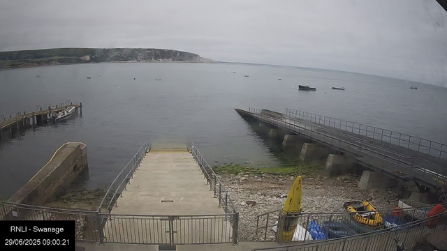 A view of a harbor with a concrete pier extending into calm water, surrounded by smaller boats and equipment. In the background, there are cliffs under a cloudy sky. Some green moss and rocks are visible near the shoreline. There are safety equipment and a small yellow vehicle at the bottom right. The timestamp shows the date and time as 29/06/2025 09:00:21.