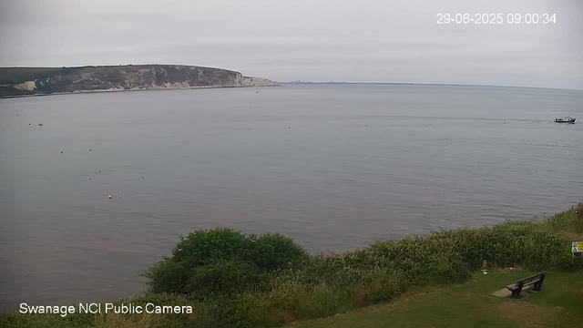 A calm body of water with a distant shoreline featuring white chalk cliffs. The sky is overcast with gray clouds. In the foreground, there is a grassy area with bushes, a bench, and a small sign. A boat is visible on the right side of the water, moving across the scene. The timestamp in the upper right corner indicates the date and time as June 29, 2025, at 9:00 AM.